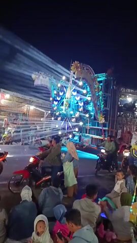 This is a vertical video taken at night, capturing a lively outdoor gathering. A large, elaborate stage structure with numerous lights and decorations, resembling a dragon's head and neck, dominates the center of the frame. People are gathered around the stage, some sitting on the ground in the foreground, others on motorcycles or standing. The atmosphere appears festive, with bright lights illuminating the scene and a crowd of onlookers. The lighting is dynamic, with beams of light sweeping across the sky and the stage, creating a theatrical effect. Several motorcycles are parked and in use, indicating transportation is a significant element of the event. Some individuals are holding mobile phones, likely recording or documenting the event. The overall impression is of a public event or celebration, possibly a concert or festival, with a focus on visual spectacle.