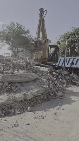 An excavator is shown loading debris into a dump truck. The excavator is yellow and has a long arm with a bucket. The dump truck is blue and has a trailer with four wheels. The scene is a construction site with piles of rubble and dirt. There are trees and buildings in the background. There are two people visible in the video. One person is operating the excavator, and the other person is standing on top of the dump truck. The video is shot from a low angle, and the sun is shining.