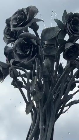 The video shows a close-up shot of a dark metal sculpture of a bouquet of roses with water spraying from the top. The camera then slowly zooms out to reveal that the roses are part of a larger sculpture of a human figure that appears to be made of intertwined metal vines. The figure is depicted in a graceful, dancing pose, reaching upwards towards the roses. The sculpture is situated in a circular fountain filled with water. In the background, there is a manicured garden with a brick pathway, blooming lavender, and hedges. Beyond the garden, a large, ornate building with towers and chimneys can be seen. The sky is cloudy.
