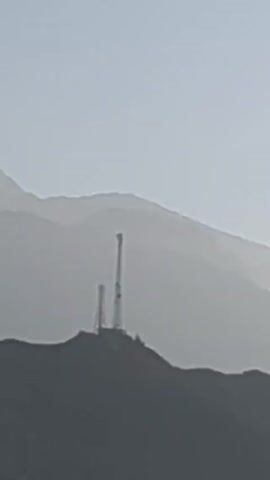The video shows a hazy, mountainous landscape with a transmission tower silhouetted on a hill in the distance. Then, a view from a window shows the landscape in daylight. A small, white pickup truck with a cage-like structure in the back is parked, and then a larger, white truck full of what appears to be coiled netting drives by. The sky is clear and blue, and the sun is visible in the background.