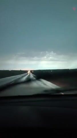 The video shows a view from inside a car driving on a wet highway during a storm. The windshield wipers are visible and appear to be in motion, clearing the rain. The sky is dark and cloudy, with some distant lights visible on the horizon. The road is reflective from the rain. The overall atmosphere is gloomy and suggests a challenging driving condition.