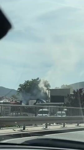 The video captures a view from inside a vehicle, looking out at an outdoor scene on a sunny day. A body of water, possibly a harbor or marina, is visible in the background, along with a boat and several large containers stacked on top of each other. Smoke is emanating from the top of one of the containers, creating a plume against the sky. The scene appears to be an industrial or commercial area, with a highway barrier in the foreground. The camera pans slightly, providing glimpses of a car and a white van.