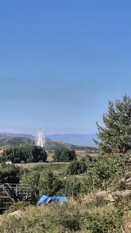 The video is a panning shot of a rural landscape under a clear blue sky. In the distance, a Ferris wheel is visible on a hilltop, surrounded by trees and mountains. In the foreground, there are trees, bushes, and some makeshift structures covered with blue tarps. The camera pans from left to right, offering a view of the diverse terrain and the distant amusement park feature.