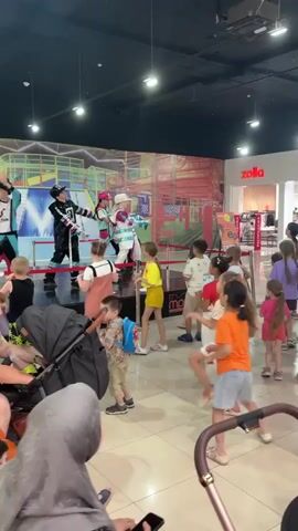 A group of children and adults are gathered in what appears to be a shopping mall, watching performers on a stage. The stage is decorated with a colorful mural of a playground or obstacle course. Three performers are on stage, dressed in costumes. The performer on the left wears a black and silver outfit with a TikTok logo. The middle performer is dressed in white with blue and red accents, holding a microphone and a blue object. The performer on the right is also in a white outfit with blue and red accents, wearing a hat and holding a similar blue object. Several children are in the foreground, some with parents, watching the performance. One performer is gesturing towards the audience. The overall atmosphere seems to be festive and engaging.