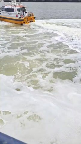 A small, white and orange boat labeled “PILOTS” is churning through the water, leaving a frothy wake behind it. The water itself is murky, with visible green and brown hues beneath the white foam. In the background, an industrial scene unfolds. There's a large, metal structure resembling an oil rig or drilling platform, and several wind turbines are visible further away against an overcast sky. The scene is captured from a slightly elevated perspective, suggesting the camera is on another boat or a platform overlooking the water. The overall impression is one of a working harbor or industrial coastal area.