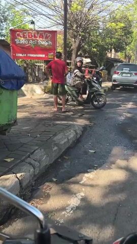 The video shows an outdoor scene on a paved street with some greenery. In the foreground, the handlebars and mirrors of a motorcycle are visible, indicating the video is shot from the perspective of a rider. To the left, a green dumpster with a blue tarp covering is present. Further down the street, a man in a red shirt and green shorts stands on the sidewalk, facing away from the camera. Beside him, a woman wearing a hijab and a patterned top is seated on a green scooter. She is looking to her left. Behind the man and woman, a red sign with white and yellow text advertising "SEBLAK BASO ACI SUGIH" is prominently displayed. There are trees and other vegetation in the background, along with a white car parked on the right side of the road. The ground appears wet in places, suggesting recent rain or washing. The lighting suggests it is daytime.