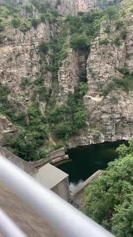 The video pans across a steep, rocky canyon filled with lush green vegetation. A concrete dam structure is visible in the foreground, holding back a dark body of water. Further into the canyon, a series of wooden platforms are suspended on the side of the cliff face, suggesting a recreational activity such as zip-lining or a viewing area. The overall environment is natural and mountainous, with a cloudy sky overhead. There are no people visible in the video.