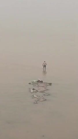 A person stands in murky water next to a small, rocky island that is littered with debris. The person is facing away from the camera and appears to be looking at something in the water. The sky is overcast, and there is a thick fog or haze in the background. The water is calm, and the overall mood of the scene is one of solitude and perhaps desolation.