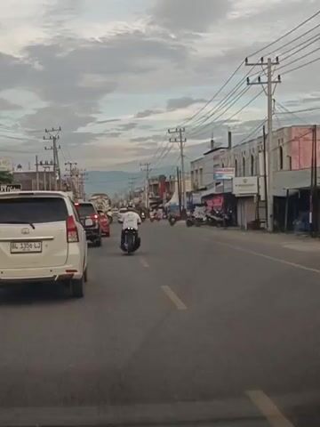 This video captures a street scene in Barat, Indonesia, likely during dusk. The perspective is from inside a vehicle, looking forward. The road is paved and has lane markings. There are several vehicles on the road, including cars and motorcycles, suggesting moderate traffic.

On the right side of the street, there are buildings lining the road, appearing to be shops or businesses. Some of these buildings have signs, though the text is largely illegible due to the resolution and distance. One sign clearly visible reads "W BARBERSHOP". Another sign above it has a logo and some text. Further down the street, more buildings and utility poles with a complex network of wires are visible.

The sky is overcast with dramatic clouds, indicating potential for rain or just a cloudy day. The lighting suggests it is either early morning or late afternoon. The overall mood is a typical urban street scene, with the activity of vehicles and pedestrians on a busy road. The presence of motorcycles is a strong indicator of the location being in Southeast Asia, and specifically Indonesia, as stated. There are no discernible emotions visible from the distant figures. The scene appears mundane and everyday, a glimpse into daily life on an Indonesian street.