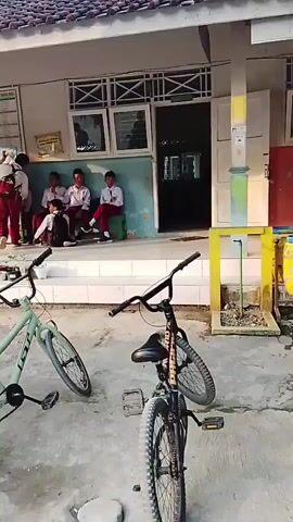 In a courtyard area with a building in the background, four children in school uniforms are seated on a low wall, with two additional children standing near them. Two bicycles are parked in the foreground, one light green and the other black. In the middle ground, a person is sitting on a motorcycle with another person behind them. The building appears to be a school or similar institution with windows and a tiled roof.