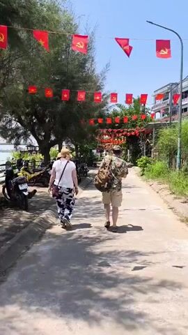 Two people are walking away from the camera down a narrow, paved road.  The person on the left is wearing a white shirt, patterned pants, and a straw hat.  They have a small bag slung over their shoulder. The person on the right is wearing a patterned shirt, khaki shorts, and a hat, and carrying a backpack.  Red flags are strung overhead across the road.  On the left side of the road are parked scooters, and a tree. On the right side of the road is vegetation.  In the distance is a white building with balconies. It is a bright, sunny day.