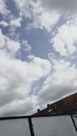 A video recorded from a low angle captures a view of a blue sky filled with white, fluffy clouds. The bottom of the frame shows the top of a white brick wall with a black trim. Above the wall, the roof of a building with terracotta tiles is visible. The clouds are moving and changing shape, with some areas of bright blue sky peeking through. The lighting suggests it is daytime, possibly with the sun filtering through the clouds.