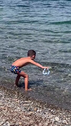 A young boy in swimming trunks is standing at the edge of the water on a pebble beach. He is holding a clear plastic bottle and appears to be pouring water from it into the sea. The water is clear and blue, and sunlight is reflecting off its surface. The boy's back is to the camera.