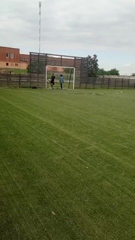 A soccer field scene with two people and a soccer ball. A green field is surrounded by a dark wooden fence. There is a soccer goal at the end of the field. A cell phone tower stands tall behind the soccer goal. One person kicks a soccer ball towards the goal, where another person attempts to block it. The sky is cloudy.