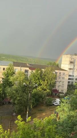 A double rainbow arcs across a gray sky over a residential area. In the foreground, lush green trees obscure the lower portions of apartment buildings. The buildings appear to be Soviet-era architecture, constructed of light-colored concrete. Some windows are visible, and balconies are present on one of the taller buildings. In the middle ground, a few parked cars are visible, including a white van. The landscape beyond the buildings appears to be rolling hills covered in vegetation. The overall impression is a post-rain scene in a somewhat dated urban environment.