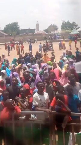 The video captures a wide shot of a large outdoor gathering in Dengi, Nigeria, during bright daytime conditions, likely under clear skies. A dense crowd of people, including many women wearing headscarves (hijabs) of various colors, men, and children, fills the foreground and midground. They are standing and looking generally forward or around, suggesting they are attending an event or celebration. Music is playing, contributing to a lively atmosphere.

In the foreground, a metal barrier or fence separates the viewer from the main crowd. Several individuals are clearly visible; a man in a white t-shirt with "YOUNG LEADERS BINGWA" written on it stands prominent, and another man wears a red t-shirt with "BULLS 23". One person in the crowd is seen carrying a white cooler on their head.

The background reveals a large, open, sandy area where more people are scattered, some standing, others moving. Tents or canopies are visible in this area, along with various buildings. A distinctive white minaret of a mosque stands tall against the sky, indicating a religious and community center. The overall scene suggests a public gathering, possibly a local festival, political rally, or community event.