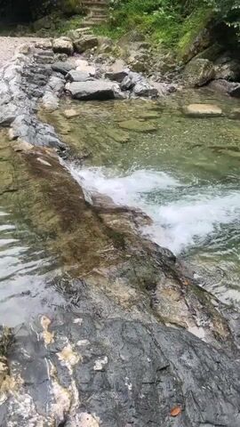 The video shows a close-up, high-angle shot of a shallow river or stream flowing over rocks. The water is clear and greenish, with white foamy rapids forming as it cascades over a ledge. The riverbed is visible, with various sizes of grey and brown rocks. The surrounding environment appears to be natural, with more rocks and some vegetation visible in the background. The lighting is natural, suggesting an outdoor setting. There are no people or animals visible in the video. The camera is stationary.