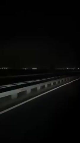 A nighttime scene viewed from a moving vehicle captures a white car driving on a dark road, likely a highway or bridge, with lights in the distance suggesting a city or settlement on the horizon. The road is marked with white lines and equipped with a barrier or guardrail. The overall setting is dark due to the night, with the only sources of light being the car's headlights and taillights, as well as the distant lights of the urban area.