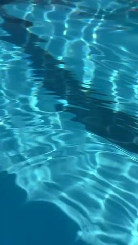The video shows an underwater view of a swimming pool. The camera pans down, revealing the blue water and the rippling light patterns on the pool floor. Lane lines are visible, marking out sections of the pool. The water is clear, and the light filtering from above creates a serene and tranquil atmosphere. There are no people or objects other than the lane lines visible in the pool.