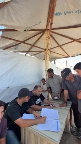 The video shows an outdoor scene under a white, umbrella-like structure with rusted metal supports. The structure is covered with a white tarp bearing the UNICEF logo. A white tent or wall is visible behind a group of people. In the foreground, two men are seated at a table covered with a striped laminate. One man, wearing glasses and a black baseball cap, is writing on a document with a pen. The other man, also seated, is looking at a small stack of cards or papers and appears to be speaking. A third man, standing behind the table, is wearing a dark grey t-shirt and a black baseball cap and is holding a small white card. He is looking down at something on the table. To his right, another man in a dark grey t-shirt and a black cap is also looking at the table. Several other individuals, including a young boy and two other men, are standing around the table, observing the activity. The ground appears to be sandy or dusty. The overall impression is one of an outdoor registration or distribution point.