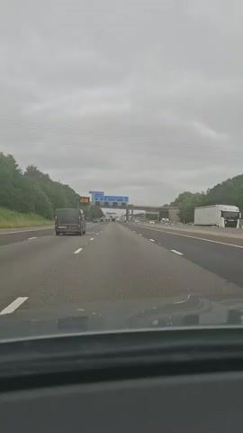 A first-person perspective video shows a car driving on a multi-lane highway under a cloudy sky. The road is paved and marked with white lines. Trees line the sides of the highway, and an overpass with large blue directional signs is visible ahead. The signs indicate directions to A653 Leeds & Dewsbury and Manchester, Bradford M62. Several cars, including a black van and a red car, are driving on the highway. The driver's side of the dashboard and windshield frame are visible at the bottom of the frame. The video appears to be captured during daylight hours.
