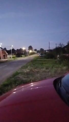 The video shows a view from a red car driving down a rural road at dusk. The sky is a deep blue with hints of pink and orange on the horizon. Streetlights are illuminated, casting a warm glow on the surroundings. On either side of the road are houses and trees. The camera is focused on the road ahead, with the hood of the car visible in the foreground. The car appears to be moving at a moderate speed. The overall impression is one of a peaceful evening drive through a quiet countryside.