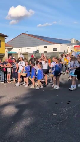 A group of young girls in matching blue dresses and white sneakers are walking in a procession down a paved street. They are accompanied by adults and appear to be part of a parade or event. The girls are mostly facing forward and some are carrying baskets or other items. The street is lined with spectators behind a metal barrier. In the background, there are buildings, a solar-powered roof, and a clear blue sky with scattered clouds. The lighting suggests it is daytime, possibly late afternoon.