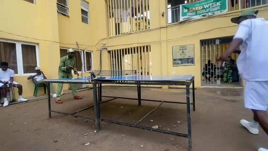 A group of young men are playing table tennis outside a yellow building. Two men are actively playing table tennis, while two other men are seated on chairs watching. One player is dressed in a green uniform and the other is dressed in white shorts and a t-shirt. The building has bars on the windows and a banner that reads "SAED WEEK". The ground is covered in dirt and debris. The weather appears to be overcast.