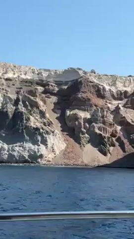 The video shows a view from a boat approaching a rocky coastline under a clear blue sky. Initially, the view features layered cliffs of varying colors – white, brown, and gray. The camera then pans to reveal a dark gray, textured cliff face rising directly from the water. A woman with dark hair wearing a bikini appears briefly in the foreground, looking at her phone while observing the cliff. The camera angle is low, indicating that it is taken from a boat moving on the water.