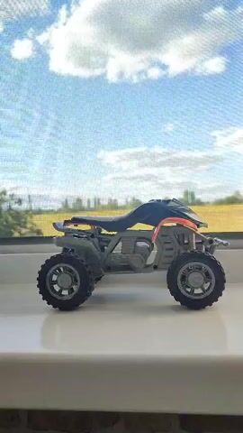 A close-up, side-view shot of a gray and blue toy ATV is positioned on a white ledge. The ATV is the primary focus, with its detailed design and features clearly visible. Behind the ATV, a window pane reveals a blurred background of trees and a bright sky with scattered clouds. The scene is well-lit, with the natural light casting a soft glow on the toy and the surroundings. The overall impression is a still-life composition, emphasizing the toy in its playful setting.