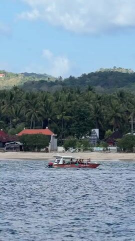 A group of people are on a red and white boat moving across the water. The boat has a white canopy and appears to be a speed boat. The people on the boat are wearing casual clothing and some are standing while others are seated. The water is blue and choppy with whitecaps. In the background, there is a sandy beach with palm trees and a few buildings. Beyond the beach, there are lush green hills covered in vegetation. The sky is blue with some white clouds. The scene appears to be a tropical location. The video is brief and shows the boat from a side angle.
