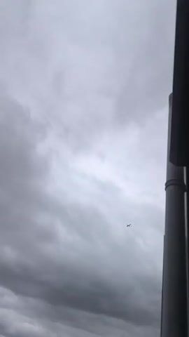 A small airplane is visible in the distance against a cloudy, grey sky. The airplane is small and appears to be flying. The sky is filled with clouds, some of which are darker than others. Part of a building or structure is visible on the right side of the frame, providing a foreground element. The overall scene is overcast and somewhat dramatic due to the cloudy sky.