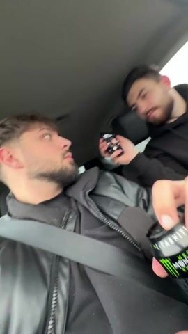 Two young men are in the front seats of a car. The driver holds a black and green can while wearing a seatbelt and a black leather jacket. The passenger in the back seat is holding a phone, taking a picture of a third man asleep in the back seat. The sleeping man is lying down, covered with a patterned jacket or blanket. The car appears to be moving on a road.