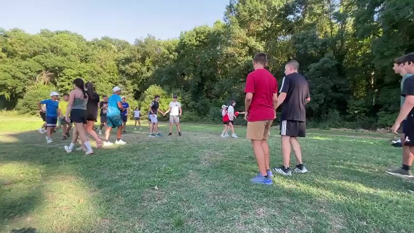 A group of children and young adults are gathered on a grassy field surrounded by trees. The weather appears to be sunny with a clear blue sky. The group is engaged in some form of outdoor activity, possibly a game or training exercise. Some individuals are interacting with each other, while others are observing or preparing to participate. The scene is dynamic, with people moving around the field. The overall atmosphere suggests a casual and energetic gathering in a natural setting.