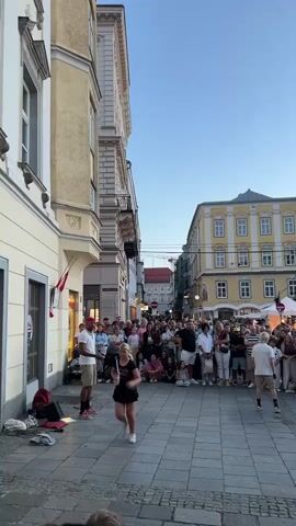 The video shows a street performance in a European town square. Two men, one wearing a red baseball cap and the other a white t-shirt, are dancing in the center of the square. A crowd of people is gathered around them, watching the performance. The buildings on either side of the square are old and ornate, with balconies and large windows. The sky is clear and blue, and the sun appears to be setting, casting a warm glow on the scene. There are some speakers and bags on the ground in the foreground, possibly belonging to the performers. The overall atmosphere is lively and festive, with people enjoying the entertainment.