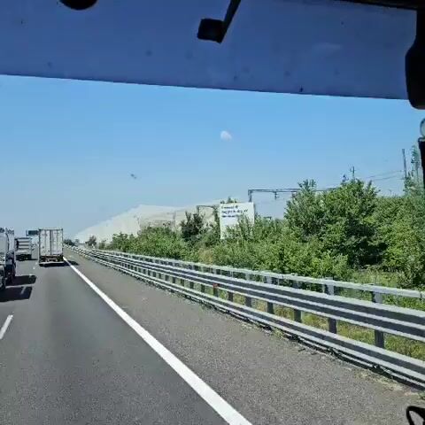 The video shows a view from the driver's seat of a vehicle moving along a highway. The sky is clear and blue. The road is asphalt with a white line separating lanes. A metal guardrail runs along the right side of the road.  There is some green vegetation growing beyond the guardrail. In the distance, a large structure covered in white material is visible. A truck is in the same lane as the camera vehicle, ahead and to the left. There is a sign in front of the structure with a caution symbol on it. Some insects are visible flying in front of the camera. The overall impression is that of a sunny day on a highway, with a glimpse of an unusual covered structure.