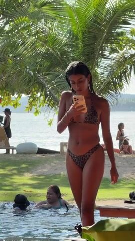 A young woman in a leopard-print bikini stands by a pool, looking at her smartphone. She has dark hair slicked back and is wearing jewelry. People are swimming in the pool, and others are walking by. Palm trees and other greenery surround the pool, and a beach and the sea are visible in the background. The scene appears to be a sunny day at a tropical resort.