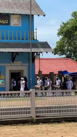 The video shows a blue building with a dark roof, likely a temple or shrine, with signage in an unreadable script. In front of the building, a white fence separates a dirt path from a paved area. Several people are gathered, some in white clothing, walking and playing musical instruments that appear to be drums. A decorative structure, possibly a fountain or altar, is visible to the left of the entrance. The environment suggests a sunny day with some trees in the background.