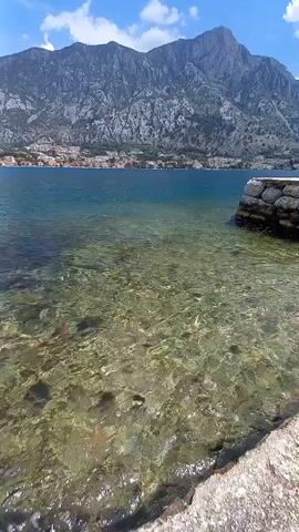 A video shows a scenic view of a body of water with a stone pier in the foreground. In the background, a town is nestled at the base of a large, imposing mountain range. The water is clear, allowing visibility of the rocky and vegetated bottom. The sky above is blue with scattered clouds. The camera is positioned at water level, looking out towards the town and mountains.