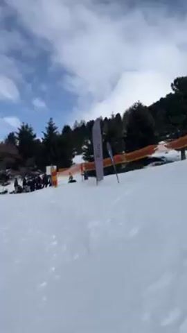 The video shows a snowy ski slope with several skiers and snowboarders, some of whom are attempting tricks on rails. The weather is overcast with a mix of blue and gray skies. There are trees lining the sides of the slope and mountains visible in the distance. Orange safety netting runs along the edge of the ski area. Various sponsor banners are placed around the slope.