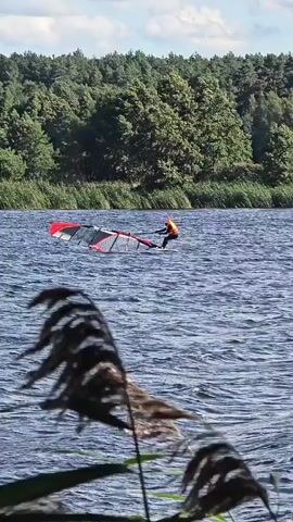 A person in an orange vest is windsurfing on a lake. The windsurfer is standing on a board and holding onto a sail. The sail is red and black. The lake is blue and choppy. There are trees in the background. The sky is cloudy.