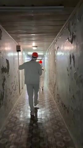 A person wearing white clothing and an orange hard hat walks down a narrow hallway. The walls of the hallway are painted white with large sections of peeling paint. The floor is tiled with a patterned design in shades of gray and white. Fluorescent lights are visible on the ceiling. Another person, also wearing white clothing, is visible further down the hallway. The person in the foreground is walking away from the camera and appears to be holding something in their right hand. The hallway has a slightly grimy and aged appearance.