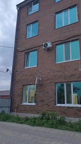 The video shows a brick building with multiple windows and air conditioning units. In front of the building, there is a paved area with some greenery. Several people are visible, including children playing with a ball and a boy sitting on a bench. A scooter is also present on the paved area. The sky is cloudy.