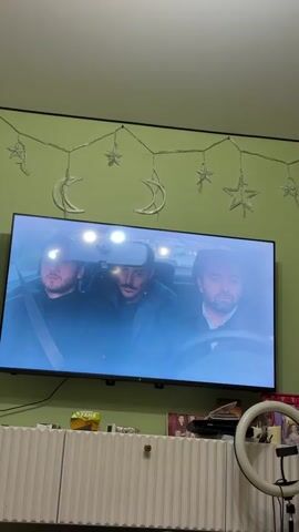Three men are in a car. The man in the driver's seat is wearing a white shirt. The man in the middle has a mustache and is wearing a dark jacket. The man in the passenger seat is also wearing a dark jacket and has a beard. The television screen is reflecting light. There are moon and star decorations on the wall behind the television. There is a white ribbed cabinet below the television. There is a ring light and a phone on the cabinet.