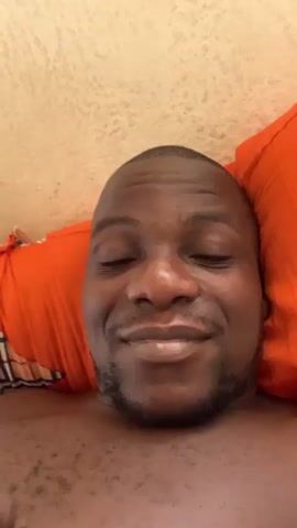 A man is lying down, possibly in bed, with an orange pillow behind his head. He is looking at the camera and smiling, holding up his hand with two fingers extended in a peace sign. The background appears to be a textured wall.