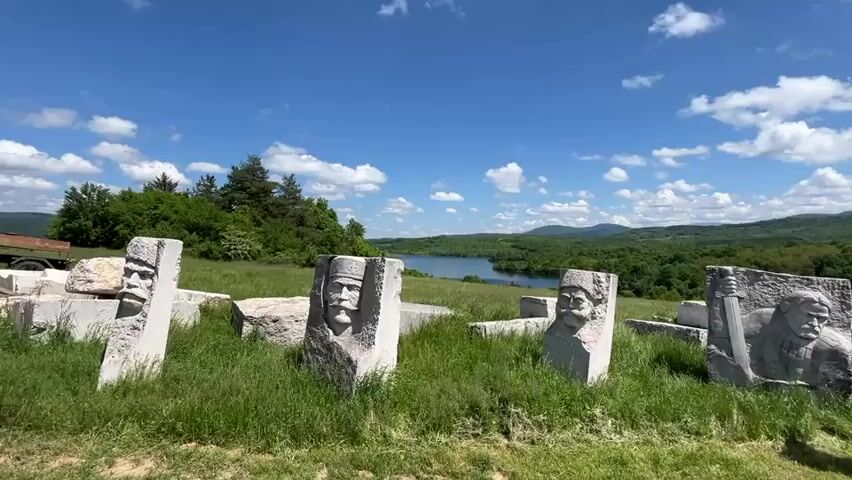 The video shows a field with several stone sculptures of heads, likely representing historical figures.  Three men are standing in the field, conversing. In the background, there is a lake, surrounded by trees and hills, under a blue sky with scattered white clouds. Some trees and a road are also visible. The day appears sunny.