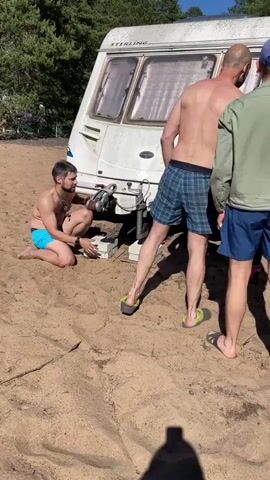 Four men are gathered around a dirty white caravan parked on a sandy surface. Trees are visible in the background. The man on the left, wearing blue swimming trunks, crouches down near the front of the caravan, holding a small object, possibly a dog, in his hands. Two other men, one in plaid shorts and the other in a green jacket and blue shorts, stand behind the caravan, appearing to be examining or adjusting it. A fourth man, partially visible on the right, wears a gray jacket and patterned shorts. The ground is covered in sand, and there are some scattered debris items. The caravan appears to be slightly tilted on its wheels. The lighting suggests it is daytime.