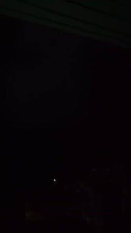 A brief video shows a dark sky with clouds. A bright flash of light, likely lightning, illuminates the clouds for a moment before the sky returns to darkness. The scene is viewed from what appears to be an interior location, with a dark ceiling or overhang visible at the top of the frame.