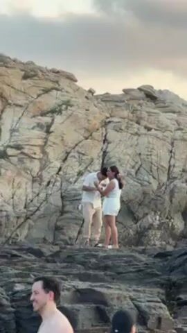 A video shows a man and woman on a rocky outcrop with a large rock face behind them. The man, wearing a white shirt and beige pants, is holding the woman's hand as they stand close together. The woman is in a white tank top and white shorts. In the foreground, other people are visible, some looking towards the couple. The environment appears to be near a body of water, possibly a beach or rocky coastline.