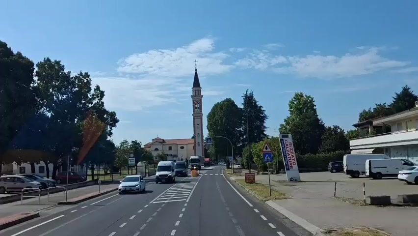 The video shows a street scene in what appears to be a European town on a sunny day. In the background, a church with a tall bell tower is prominent. A road with white lane markings runs through the center of the frame. Several vehicles are visible on the road, including cars, vans, and trucks. Pedestrian crossing signs are present, indicating a crosswalk. Trees line the sides of the road, and buildings can be seen on the right side of the frame. The sky is blue with some clouds. The camera appears to be mounted on a moving vehicle, suggesting a first-person perspective from someone traveling along the road.